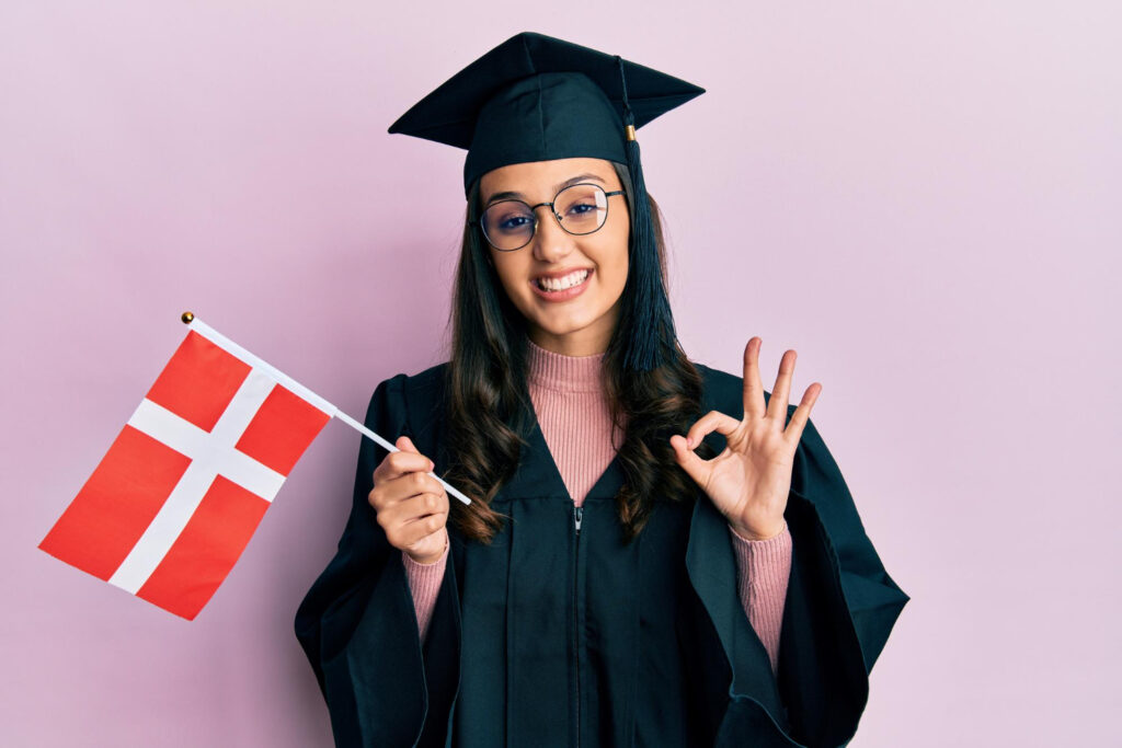 young hispanic woman wearing graduation uniform holding denmark flag doing ok sign with fingers smiling friendly gesturing excellent symbol